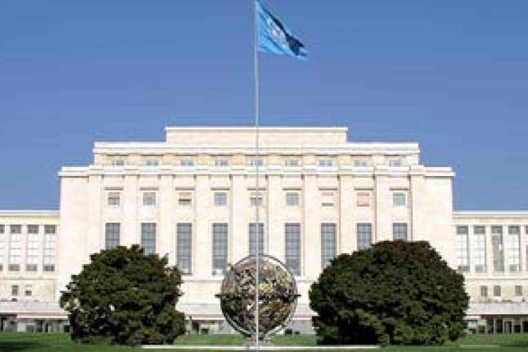 Large neoclassical building with a UN flag on top, a globe sculpture in front, and two trees on either side, set against a clear blue sky.