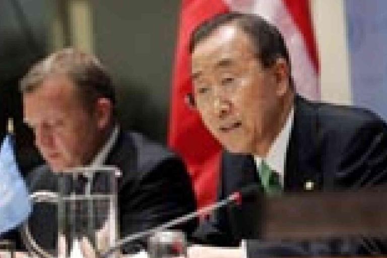 Two men in suits sit at a conference table with microphones, one speaking and one writing, with flags and documents visible in the background.