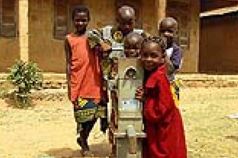 Five children stand around a manual water pump in an outdoor setting, with a building in the background.