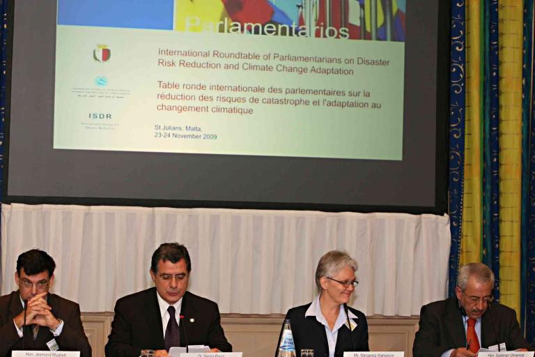 Four people sit at a table in front of a large screen displaying a presentation on disaster risk reduction and climate change adaptation at an international roundtable event.