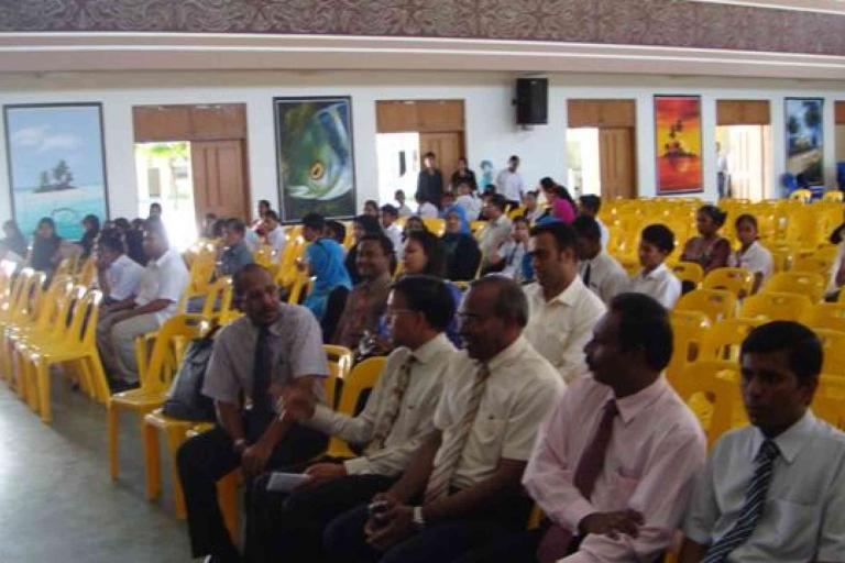 People seated on yellow chairs in a hall, with some attendees talking and others looking forward; wall art and windows are visible in the background.
