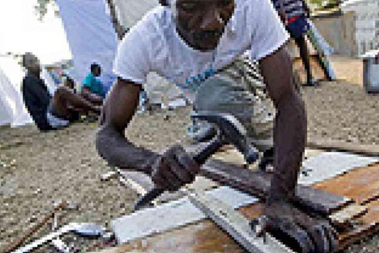 A man in a white shirt repairs wooden boards with a hammer outdoors, while others sit in the background on a gravel surface.