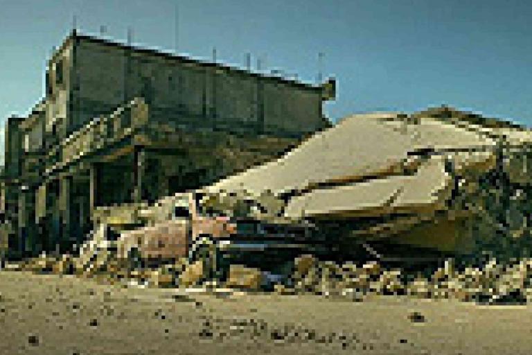 A collapsed building with debris covering a car on a street, suggesting recent earthquake damage and destruction.