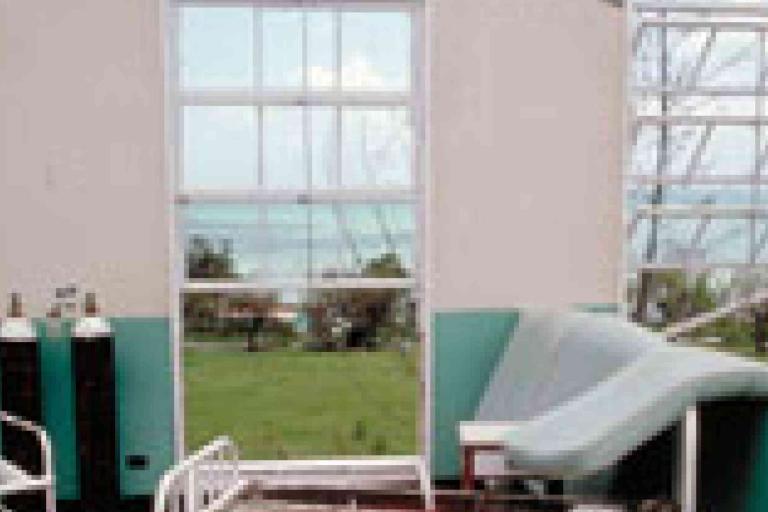 A hospital examination room with a hospital bed, oxygen tanks, and large windows overlooking a grassy area and a body of water.