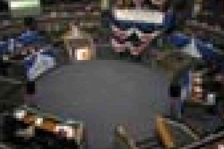 Blurry image of people seated in a circular arrangement around a central area, possibly in a meeting or conference setting.