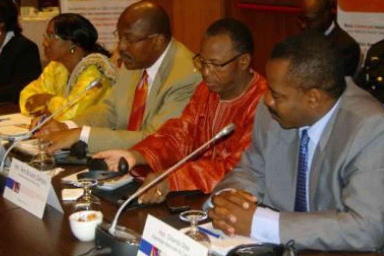 Four individuals sit at a conference table with microphones and documents, engaged in discussion at a formal meeting.