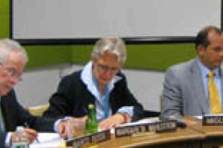 Three people in business attire sit at a table with nameplates, papers, and drinks in front of them, appearing to participate in a formal meeting.