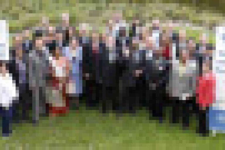 A large group of people poses outdoors on grass between two standing banners, with greenery in the background.