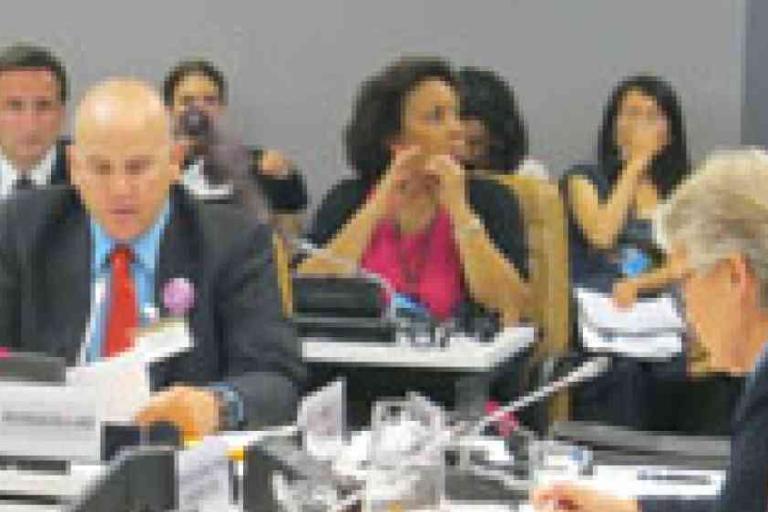 Several people sit at conference tables during a formal meeting, with microphones and documents in front of them.