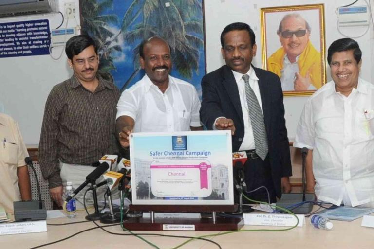 Five men stand around a table at a press event, unveiling a sign titled "Safer Chennai Campaign." Microphones and documents are on the table. A portrait hangs on the wall behind them.