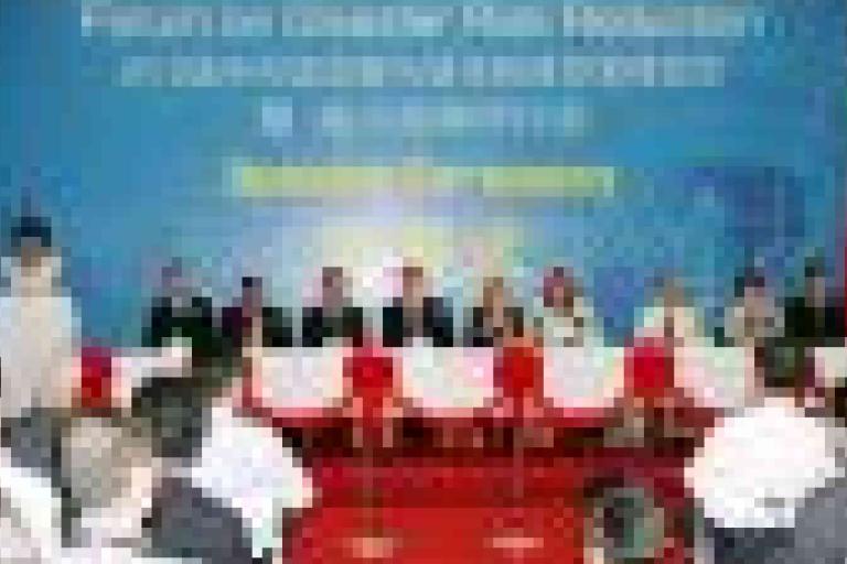 A group of people sit at a long table on a stage with a blue banner behind them at a formal event. Audience members are seated in front.