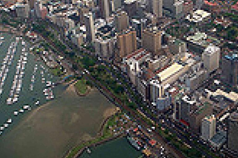 Aerial view of a coastal city with high-rise buildings, a marina filled with boats, and green spaces along the waterfront.