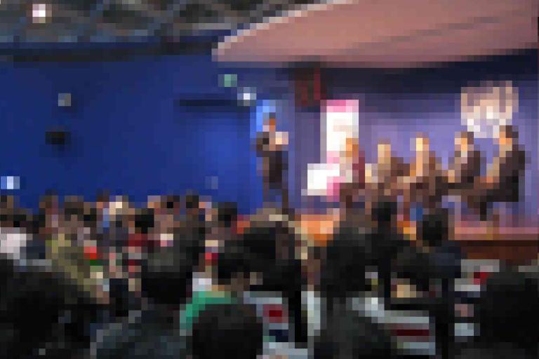 A large audience watches a panel of speakers seated on a stage in a blue-walled auditorium.