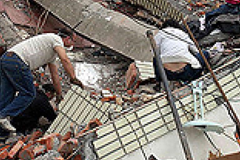Two people search through debris and rubble of a collapsed building.