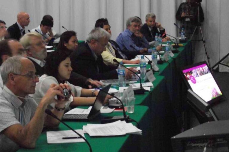 A group of people sit at tables with laptops, papers, and water bottles, attending a conference or meeting; a presentation screen is visible in the foreground.