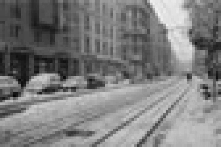 A city street in winter with snow covering parked cars, tram tracks, and sidewalks, lined with multi-story buildings and bare trees.