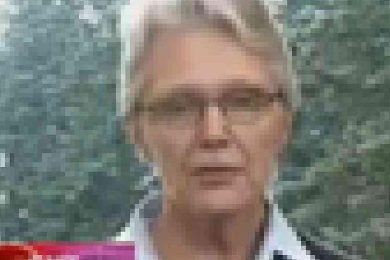 A pixelated image of a person with short light hair and glasses, standing outdoors with blurred trees in the background.