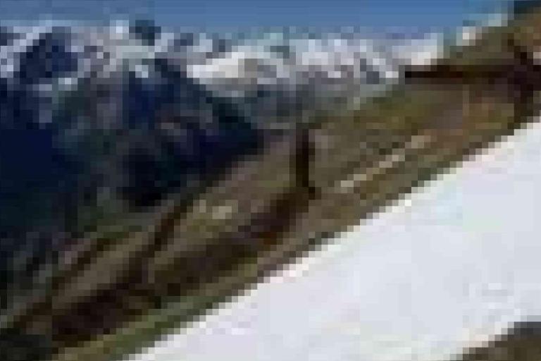 A blurry image shows a steep mountain slope partly covered with snow, with snow-capped peaks in the background under a blue sky.