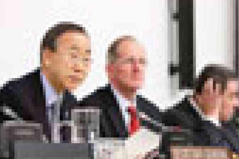Three men in suits sit at a conference table with microphones and nameplates, appearing to participate in a formal meeting or panel discussion.