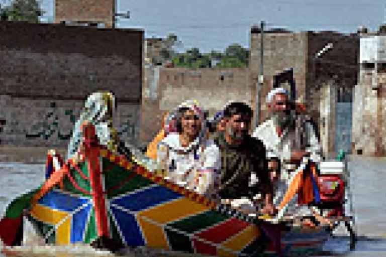Four people ride a brightly painted wooden boat through a flooded urban area, with water covering the street and buildings visible in the background.