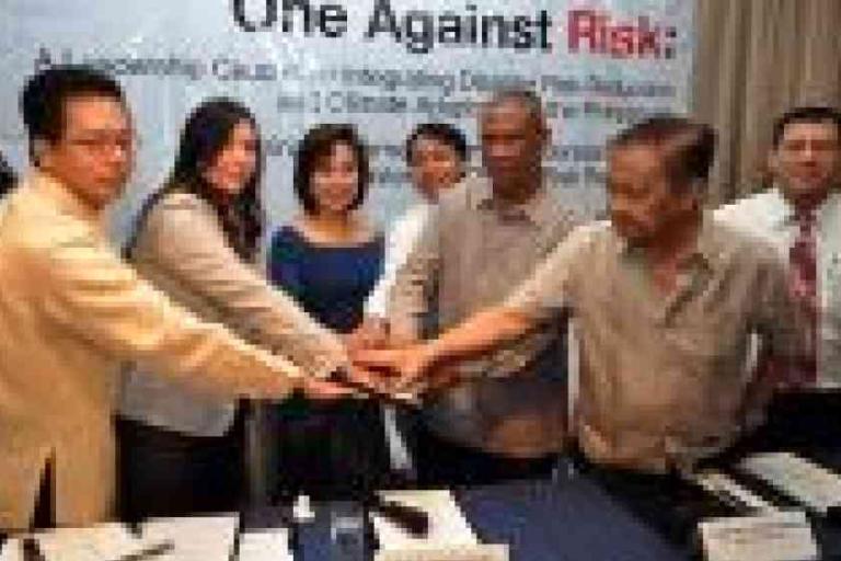 A group of six people stand together at a table, joining hands in the center, with a conference banner in the background.