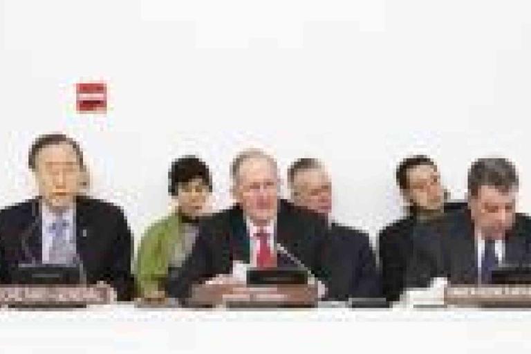 Six people sit at a conference table with microphones and nameplates in front of them, participating in a formal meeting.