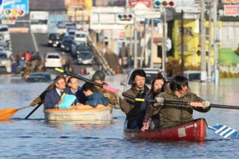 People paddle boats through a flooded city street as cars are partially submerged and traffic is halted in the background.