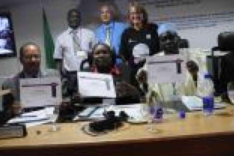 A group of people at a conference table hold certificates while others stand behind them, with documents and beverages on the table.