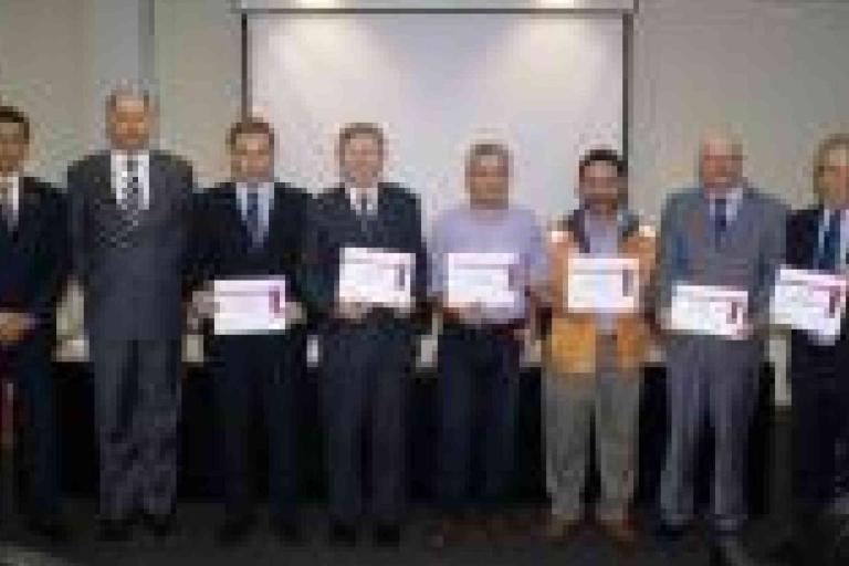 Eight men stand in a row indoors; four of them in the center hold certificates. A projection screen and flag are visible in the background.
