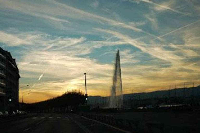 A city street at sunset with a tall water fountain in the distance and streaked clouds in the sky.