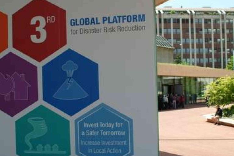 A sign for the 3rd Global Platform for Disaster Risk Reduction stands outdoors near a modern building, with icons illustrating various disaster types.