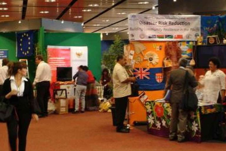 People interact at various booths during an indoor event focused on disaster risk reduction, with informational displays and banners visible.