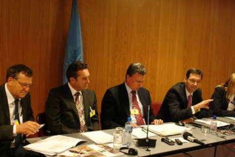 Five people in business attire sit at a conference table with documents, microphones, and water bottles in front of them, in a wood-paneled room with a blue flag in the background.