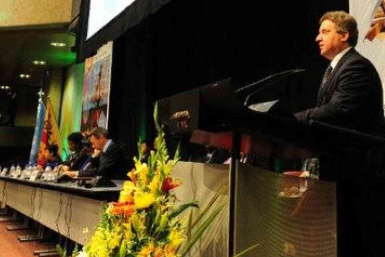 A man in a suit speaks at a podium on a stage, with several people seated at a long table beside him and flower arrangements in the foreground.