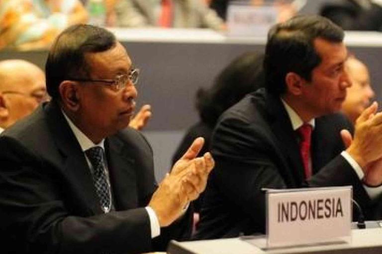Two men in suits sit and applaud at a conference table with a sign labeled "Indonesia" in front of them.