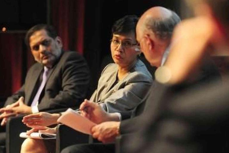 Three people in business attire sit on a panel, engaged in discussion, with one person gesturing while speaking and others listening attentively.