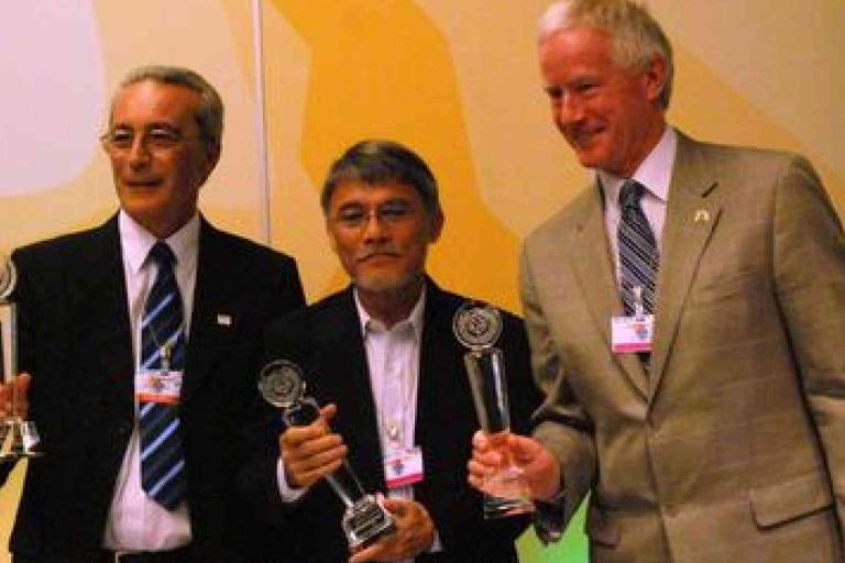 Three men in suits stand side by side, each holding a glass award and posing for a photo at an indoor event.