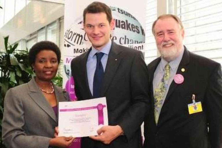 Three people in business attire pose indoors; one holds a certificate while the others stand beside him, smiling at the camera.