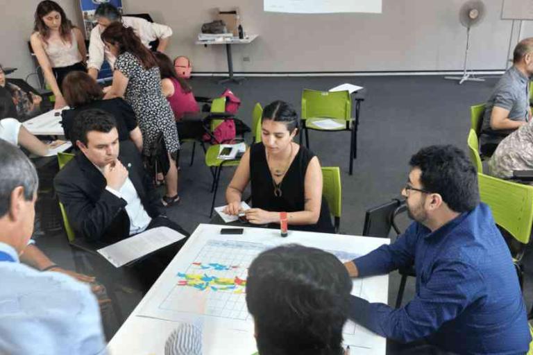 A group of people sit around tables in a classroom, engaging in discussion and paperwork; maps and charts are spread out on the tables.