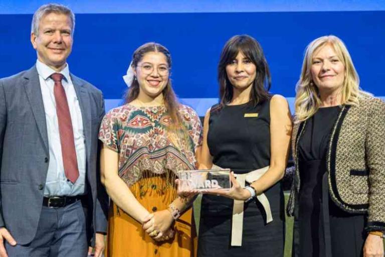 Four people stand together onstage, one holding an award plaque, all dressed in formal or semi-formal attire, posing for a group photo against a blue background.