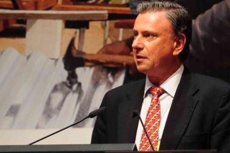 A man in a suit and patterned tie speaks at a podium with microphones, with a blurred background featuring construction materials.