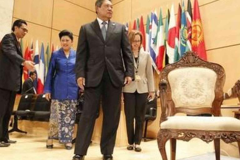 A group of formally dressed people walk past an ornate empty chair in a conference room with many international flags in the background.