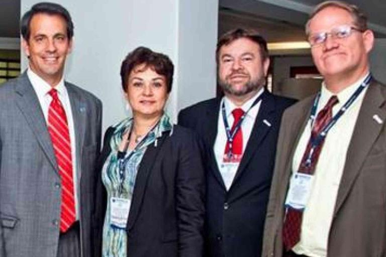 Four people in business attire stand side by side indoors, all wearing name badges and looking at the camera.