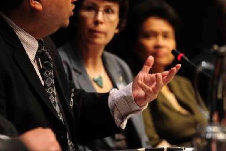 A man in a suit speaks into a microphone at a panel discussion, with two women seated beside him, listening attentively.