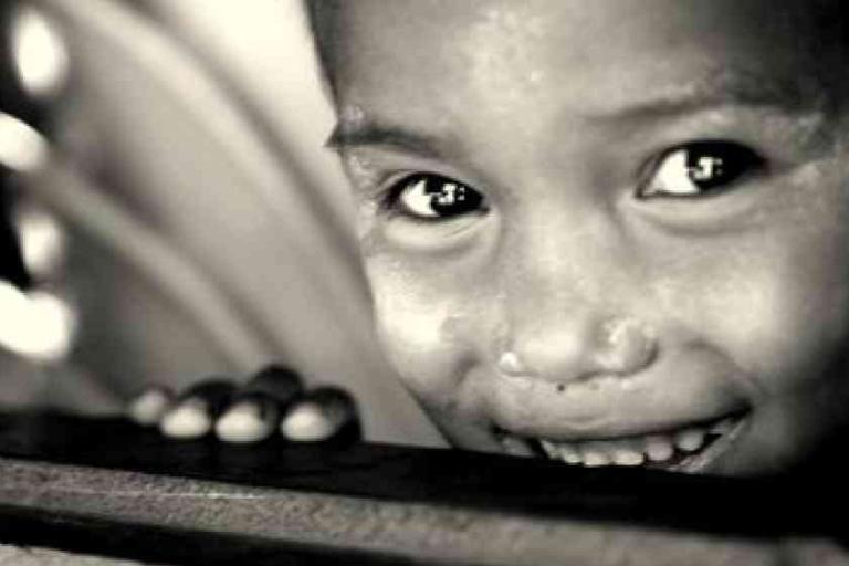 Close-up black and white photo of a smiling child with bright eyes, partially hiding behind a wooden surface.