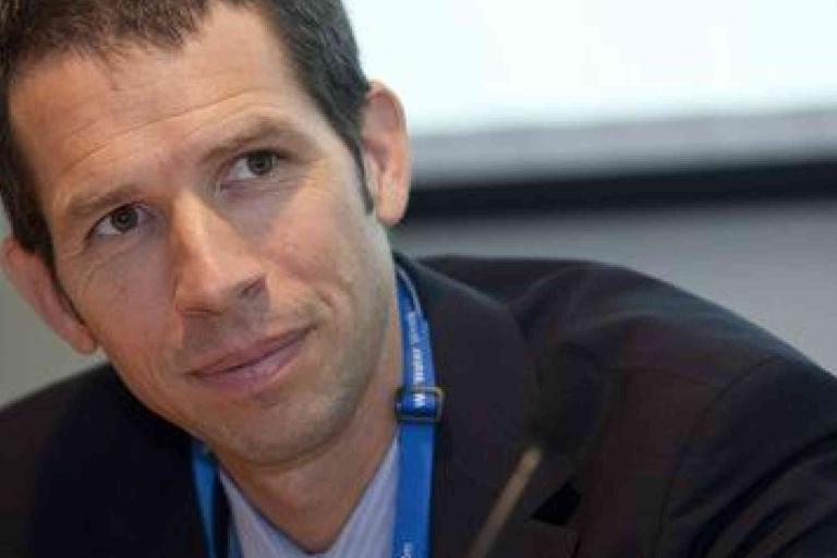 A man in a suit with a blue lanyard is looking slightly to the side and smiling during an indoor event or meeting.