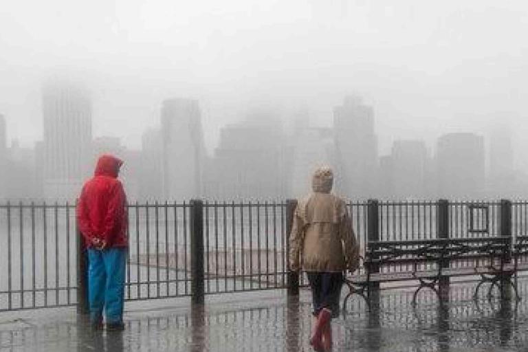 Two people in raincoats stand by a wet waterfront railing, with benches nearby and a city skyline obscured by heavy fog in the background.