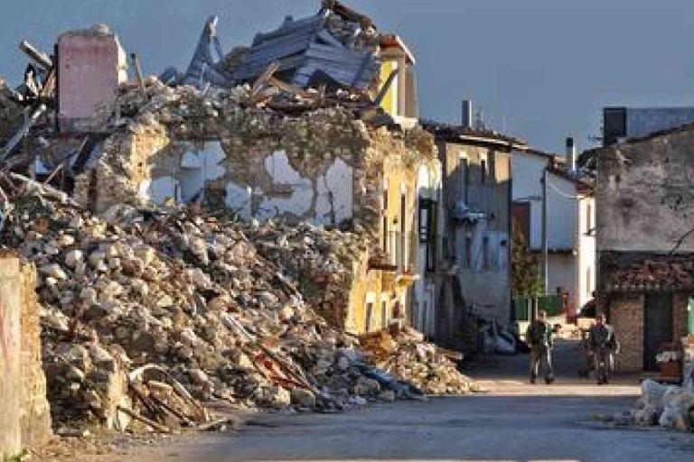 Collapsed building with rubble spilling onto a street after an earthquake; two people walk in the background near damaged structures.