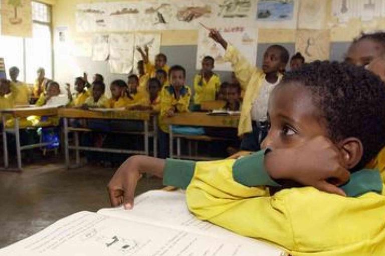 A classroom of young students wearing yellow uniforms; one student sits at the front with an open book while others look toward a peer raising their hand.