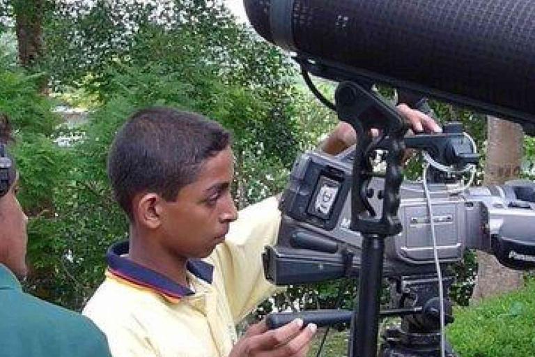 A young person adjusts a Panasonic video camera on a tripod outdoors, with another person nearby wearing headphones.